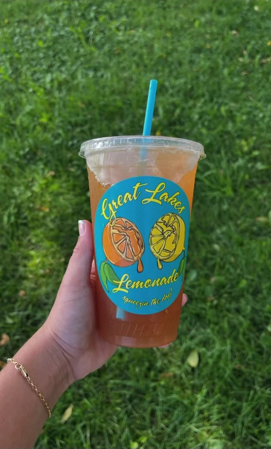 A hand holding a branded Great Lakes Lemonade cup against a green grass background