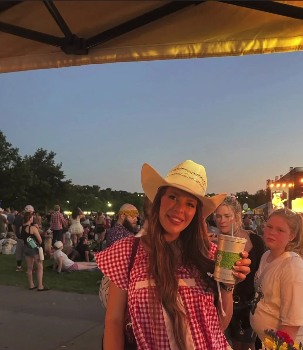 A person in a cowboy hat holding lemonade behind a flower-decorated booth in the evening