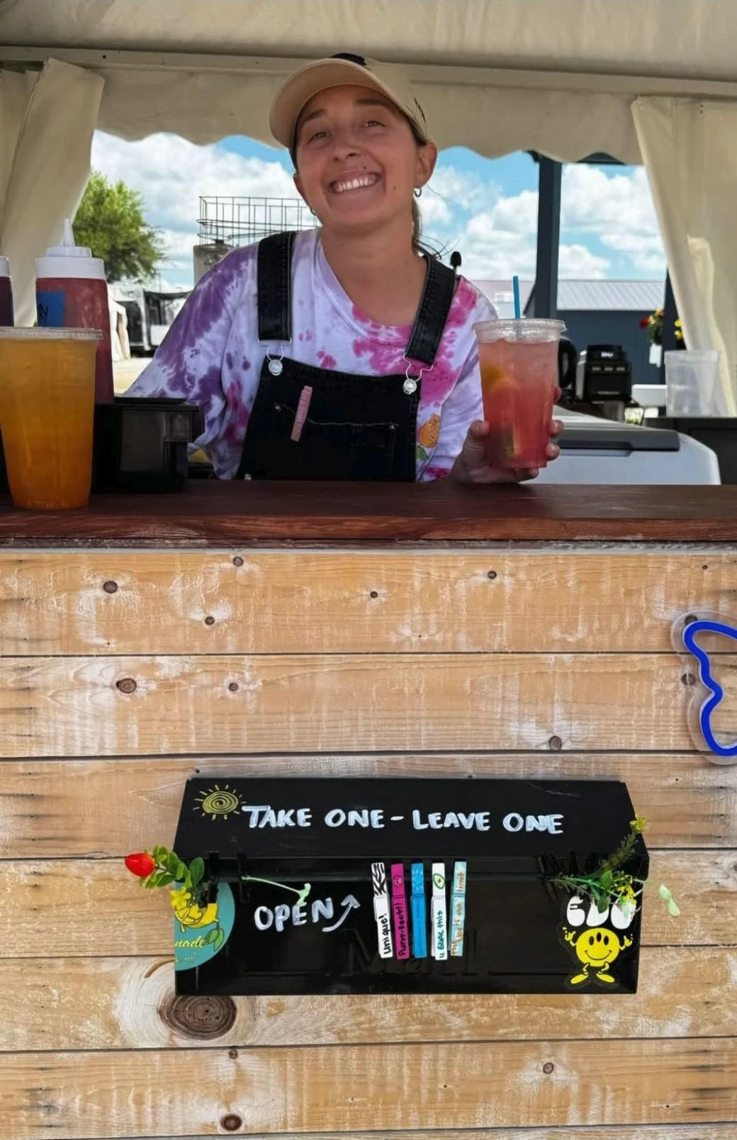 A smiling person holding a red drink behind a wooden lemonade counter