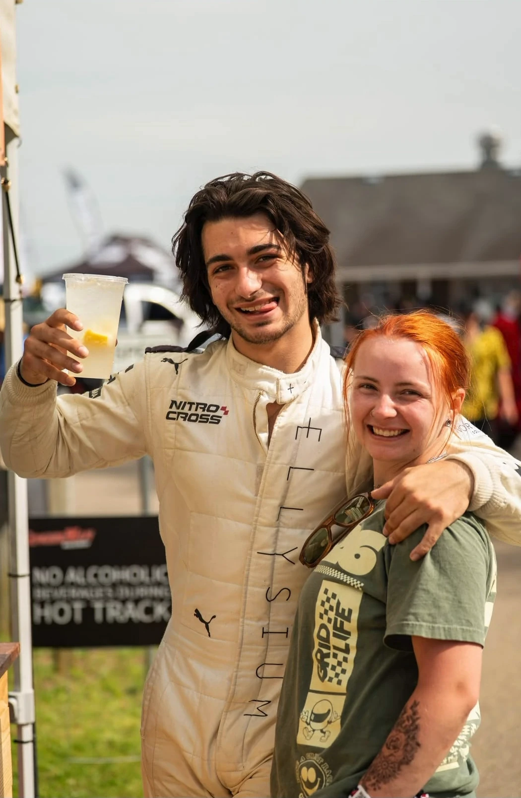 A guest in a racing suit holding lemonade beside a smiling team member at an outdoor race venue