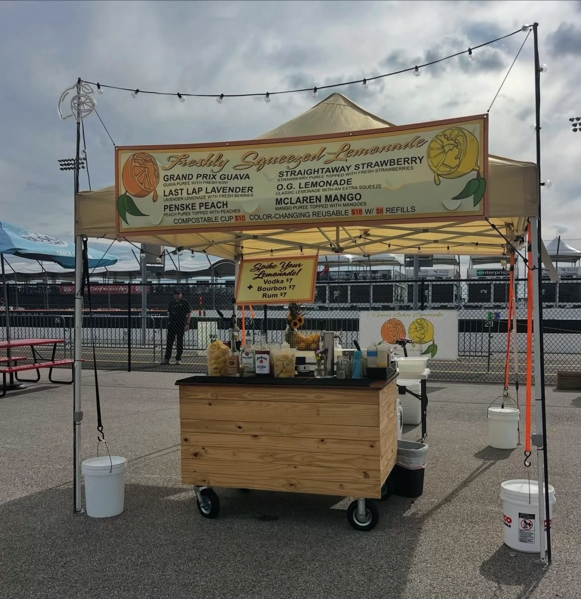 A wood-front lemonade booth and menu under a canopy beside a racetrack