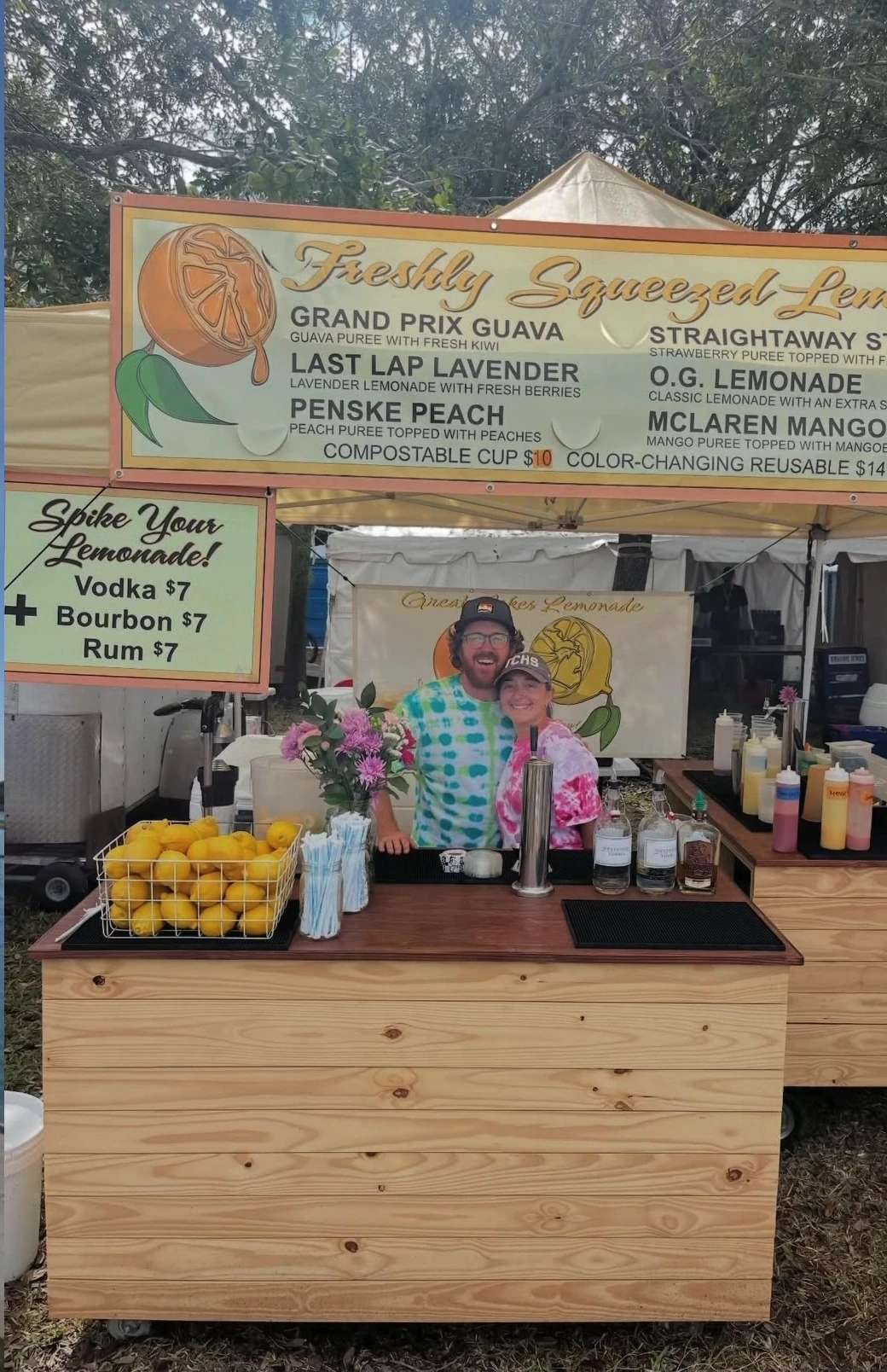 Two team members at a lemonade counter with menus, lemons, bottles, and flowers